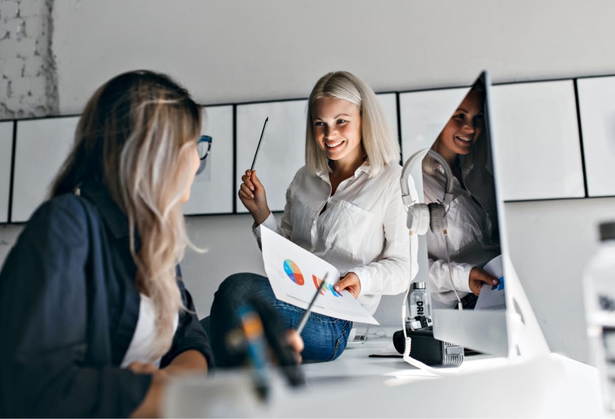 smiling-blonde-female-manager-holding-infographic-pencil-while-sitting-table-indoor-portrait-two-women-working-with-computer-office